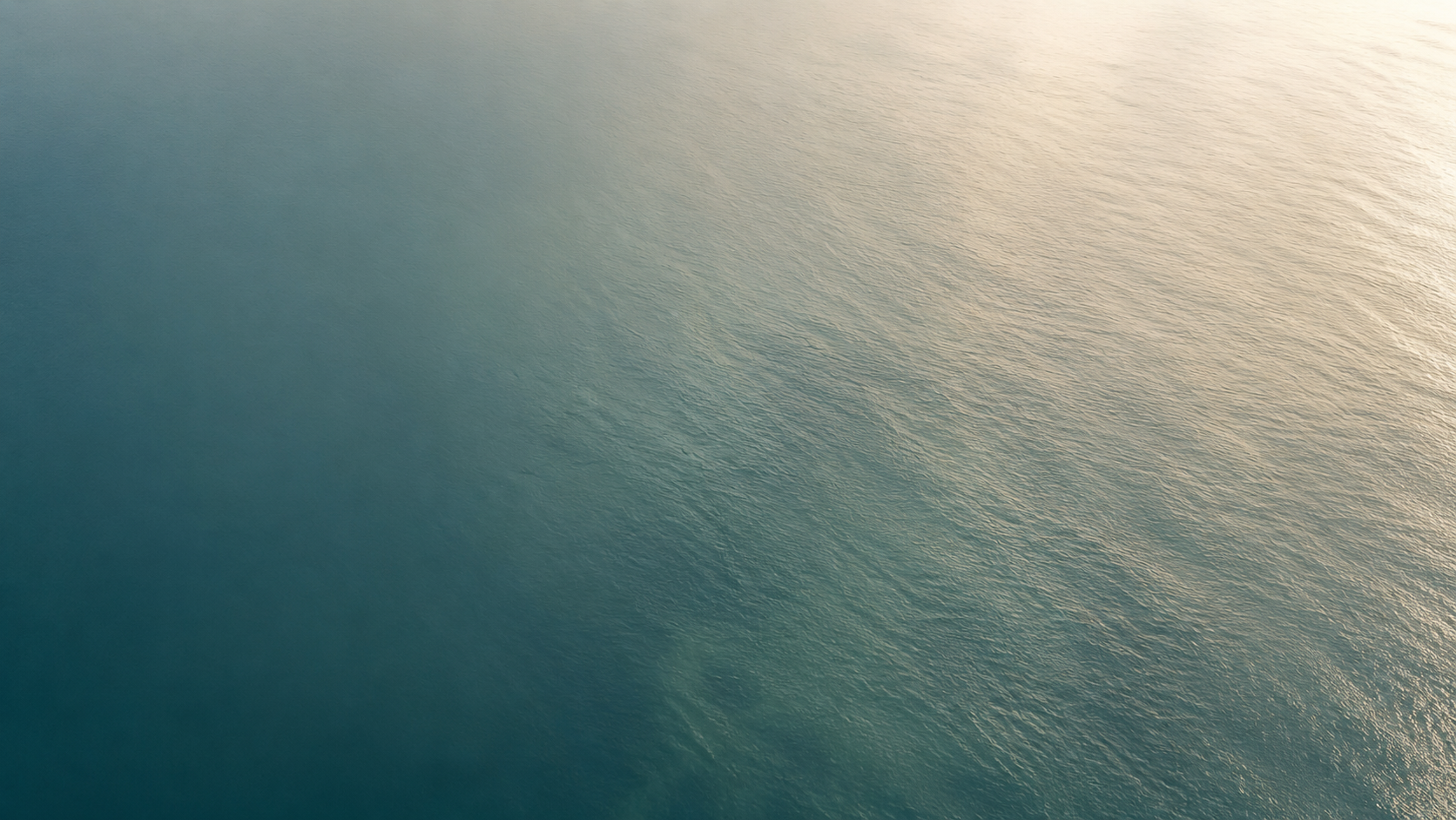 Aerial view of calm ocean water shifting from deep teal to a warm horizon